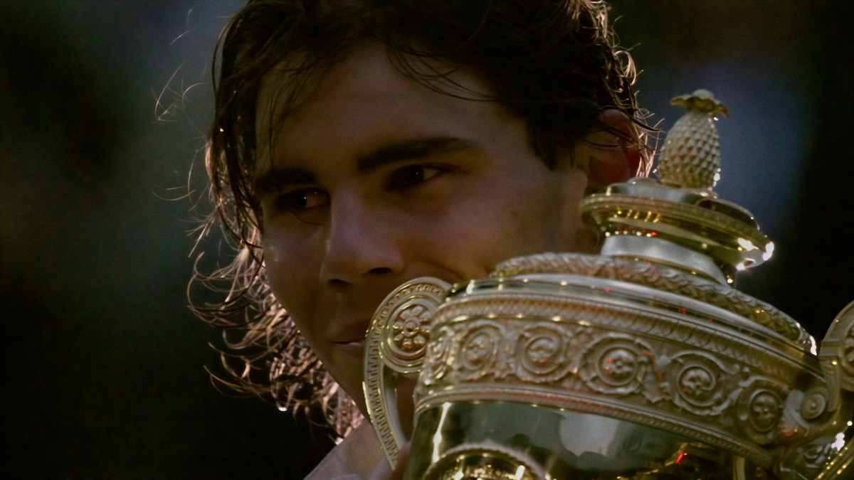 A tennis player holding a large gold trophy, partially visible face, in an outdoor setting with dramatic lighting, celebrating a victory.