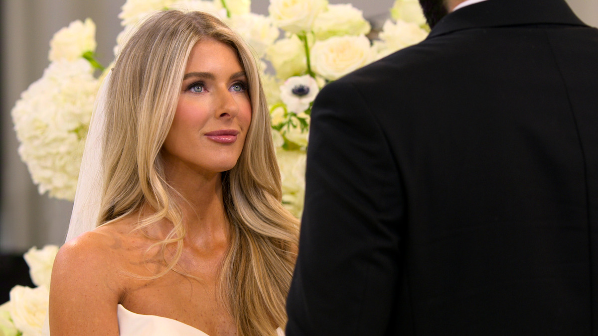 A bride in a white dress and veil stands facing a man in a black suit during a wedding ceremony, surrounded by white flowers and soft lighting.