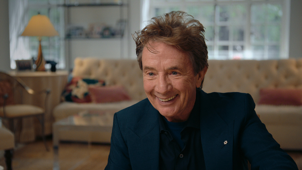 Smiling man with tousled hair in a dark blazer sits in a well-lit, cozy living room with a tufted sofa, cushions, lamp, shelves, and large windows in the background.