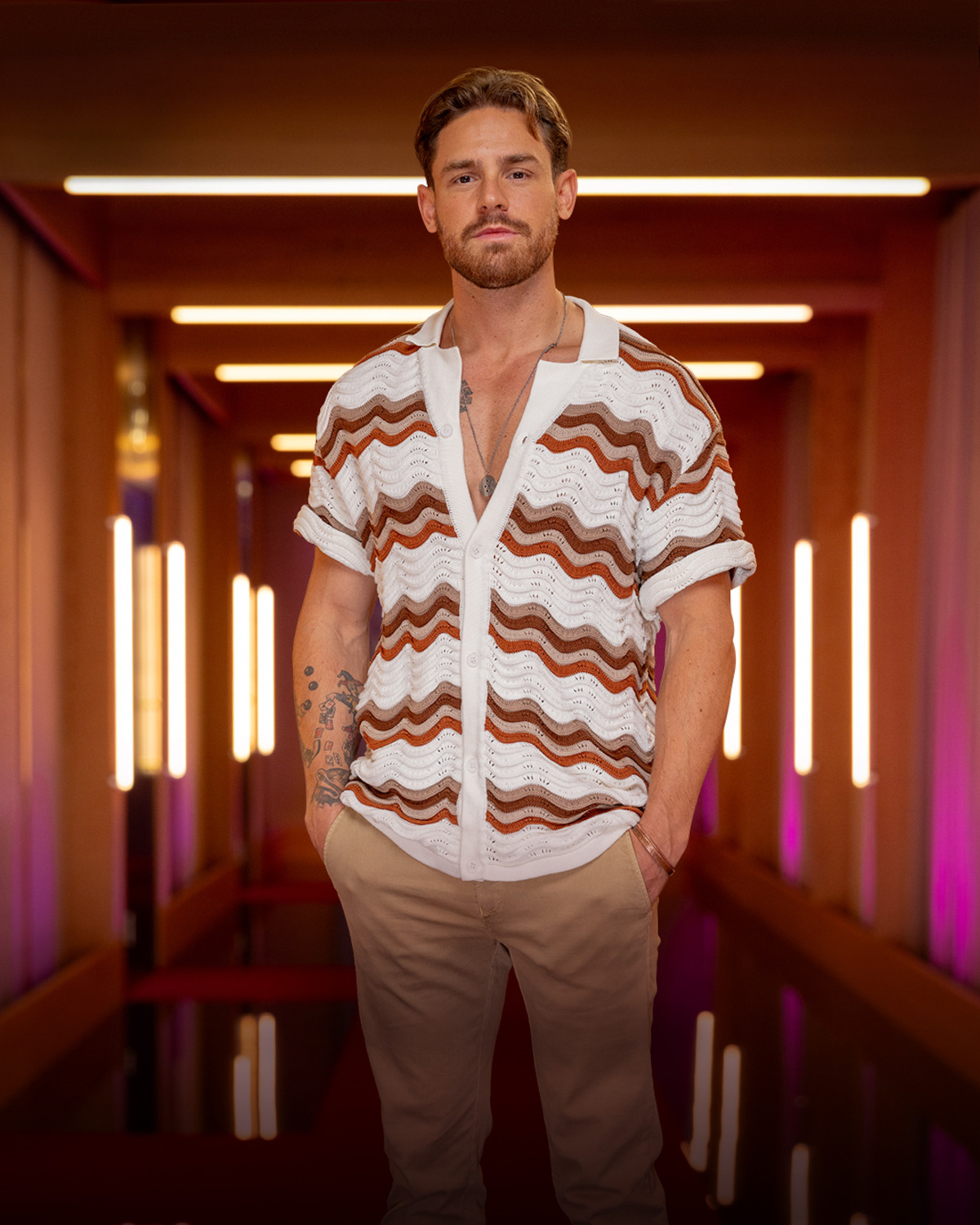 Man with light brown hair and a beard, wearing a white and brown wavy-patterned shirt and beige pants, standing in a modern hallway with warm lighting and neon accents.