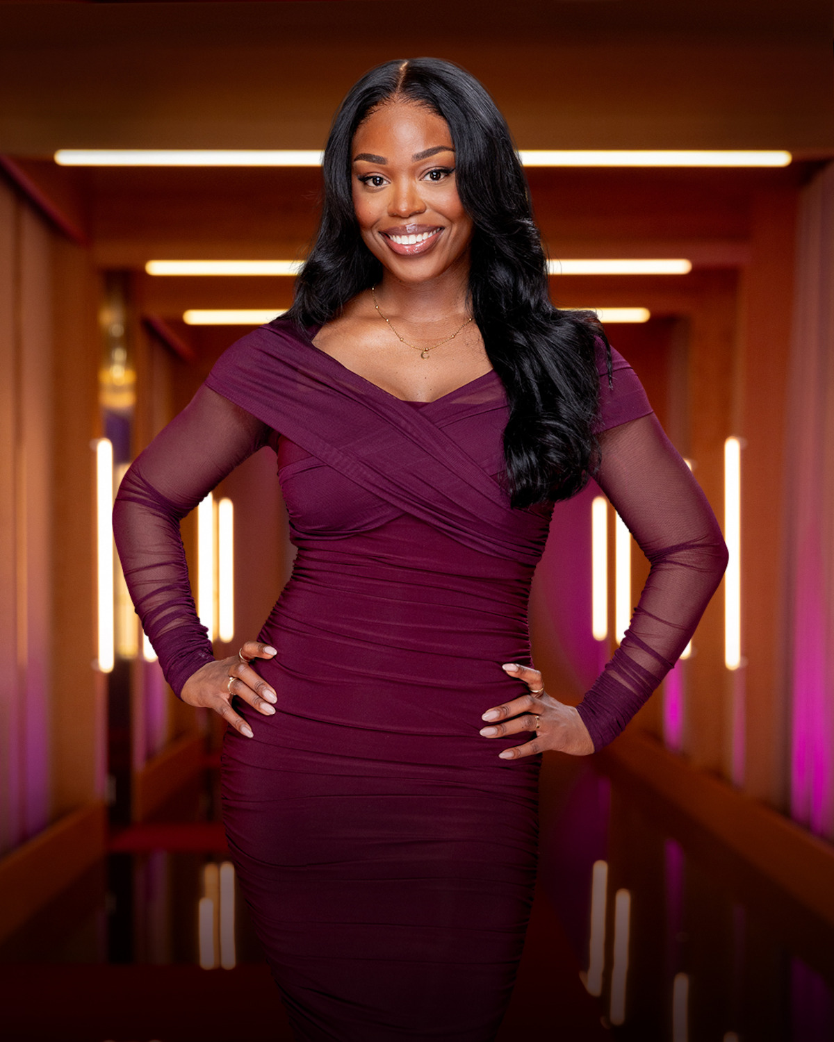 Smiling woman in a fitted, long-sleeve burgundy dress stands confidently in a modern, warmly-lit hallway with glowing light panels and soft pink accents in the background.