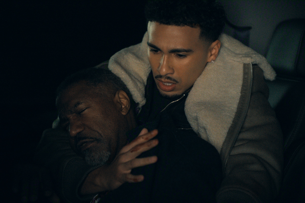 A young man in a shearling jacket cradles an older man who appears unconscious or unwell inside a dimly lit car, expressing concern and urgency in a tense setting.