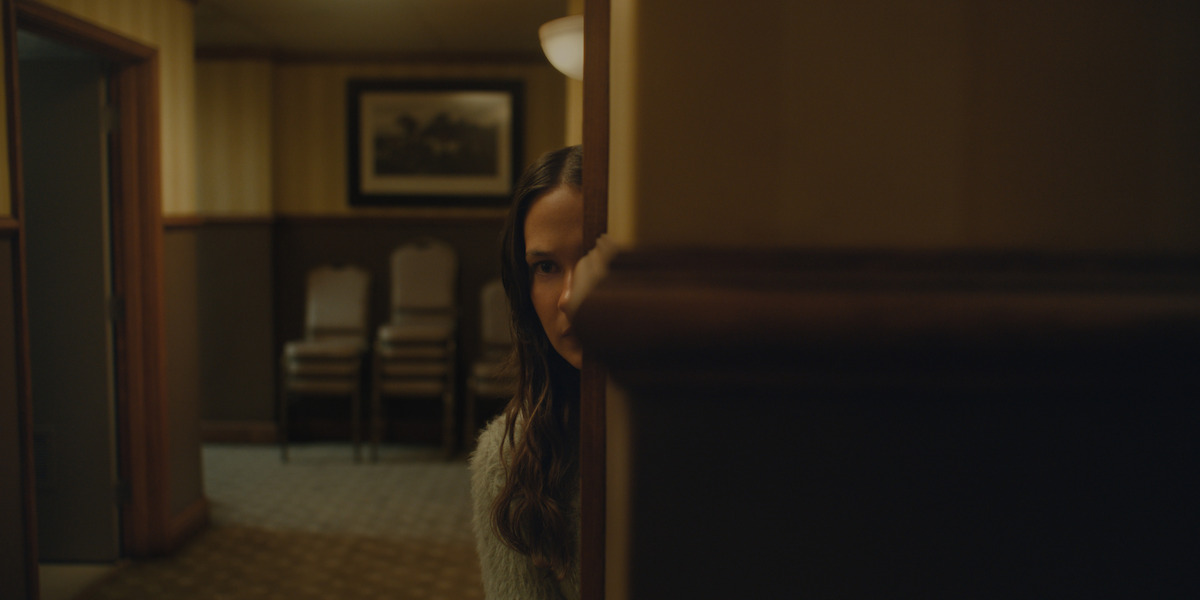 A woman partially hidden behind a wall, peering into a dimly lit room with stacked chairs, patterned carpet, and framed art, suggesting a quiet, tense atmosphere in an indoor setting.