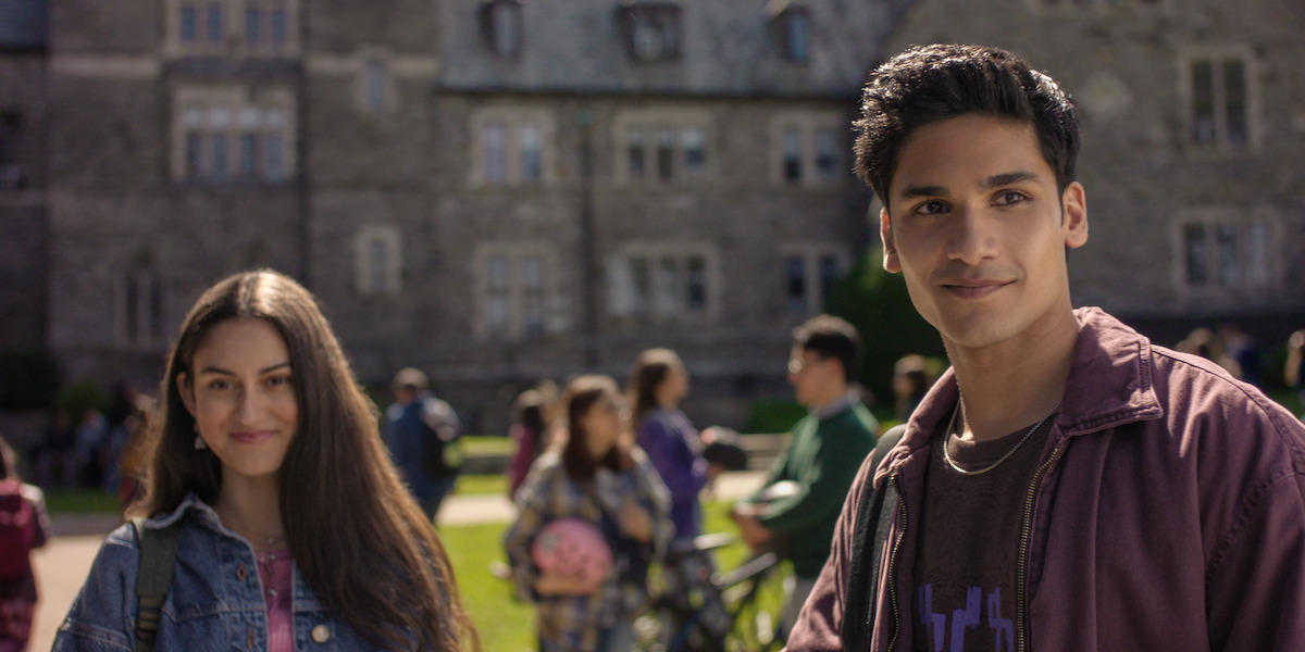 Two young people standing outside, smiling, with a college campus and other students in the background on a sunny day.
