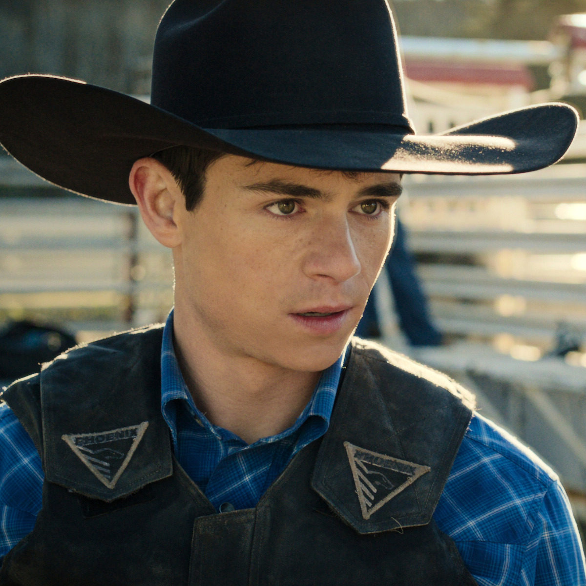 Young man in cowboy hat and plaid shirt stands outdoors at a rodeo or ranch setting, wearing a protective vest, talking to another cowboy with livestock and fencing in the background.