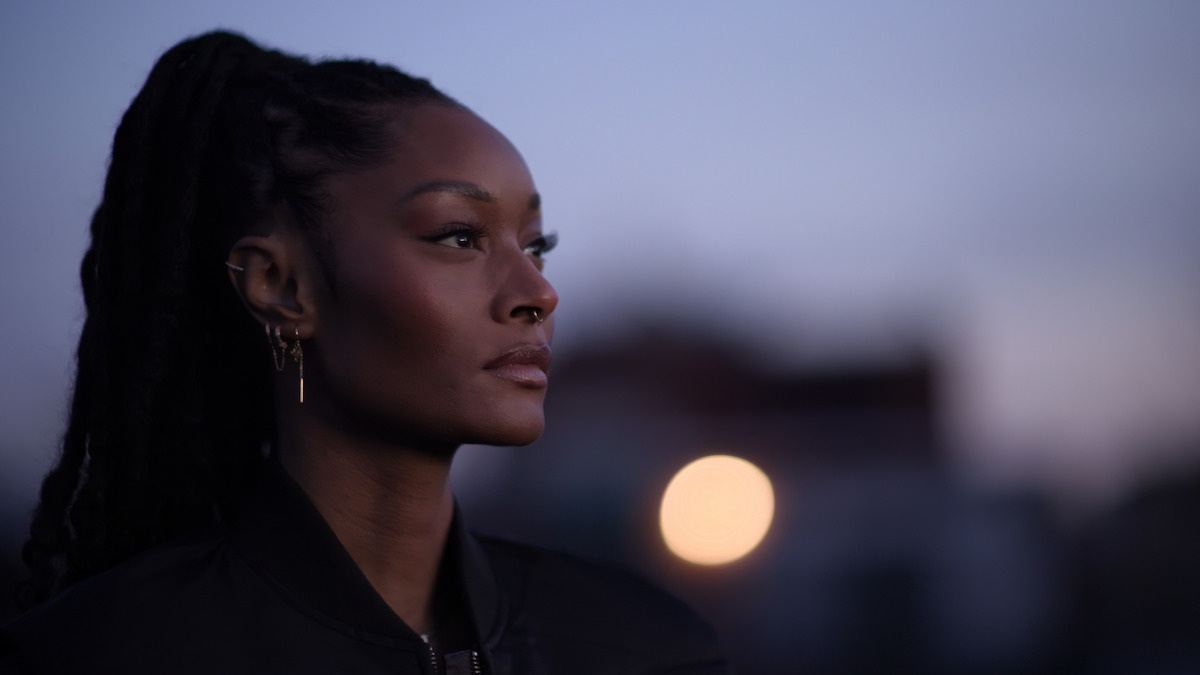 Woman with long braided hair and gold earrings gazes thoughtfully to the side outdoors at dusk, blurred urban background with soft light and a bokeh effect.