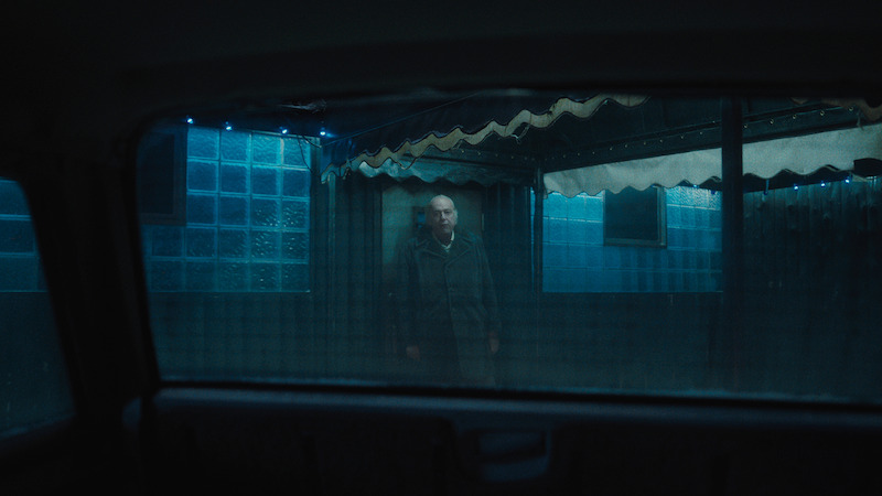 A man stands alone outside under a canopy, viewed through a car window at night. Blue lighting and tiled walls create a moody, isolated atmosphere.