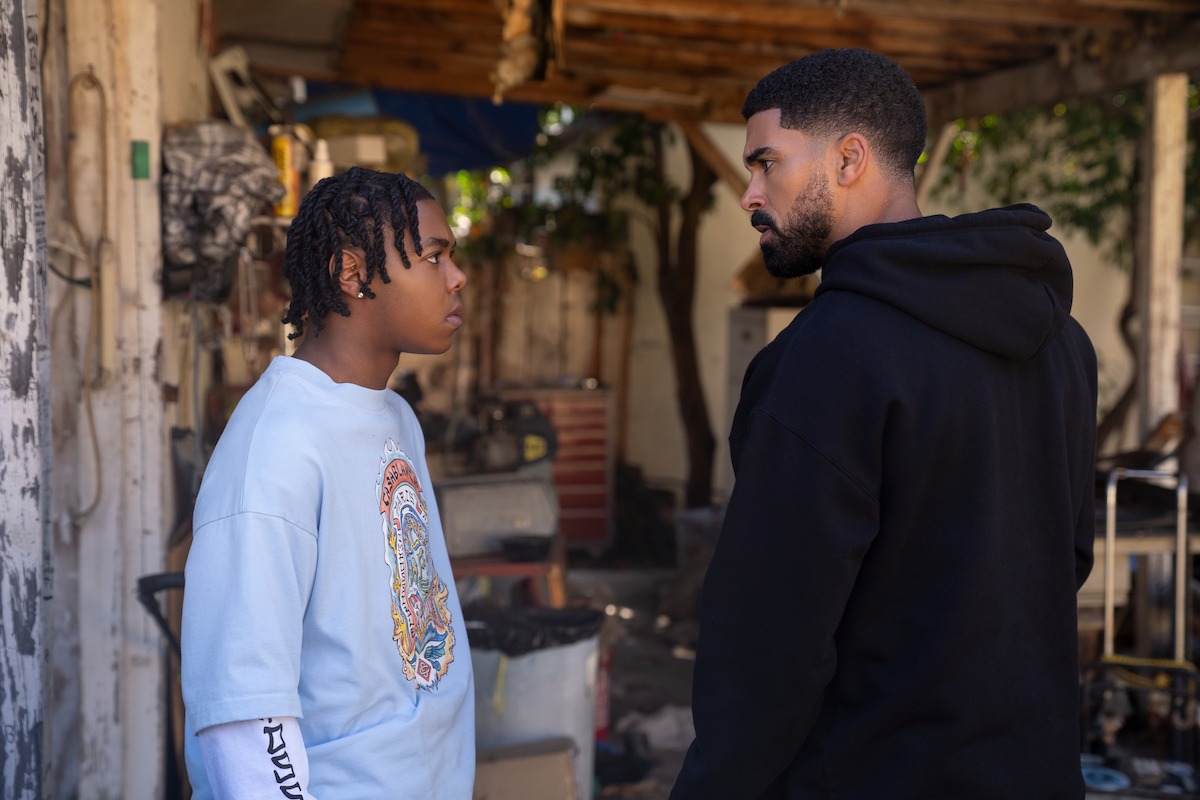 Two young men stand face-to-face in a tense moment outdoors, surrounded by a cluttered, partially covered area with scattered debris, broken walls, and trees in the background.