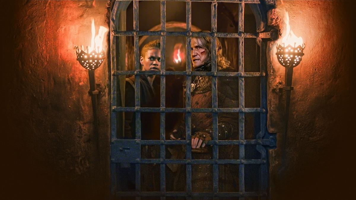 Two people stand behind iron bars, illuminated by torches on either side, creating a tense and dramatic atmosphere.
