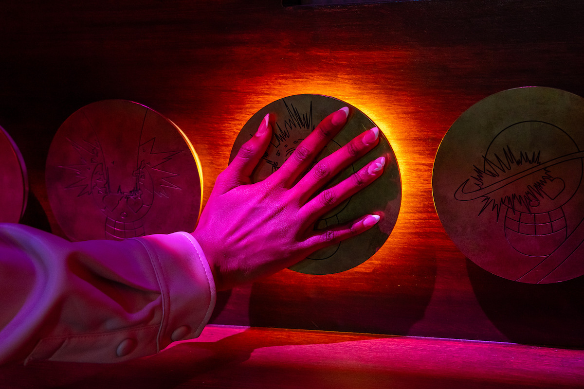 A hand with painted nails presses a round, illuminated brass panel with engravings on a wooden surface, in a dimly lit setting with dramatic pink and orange lighting.