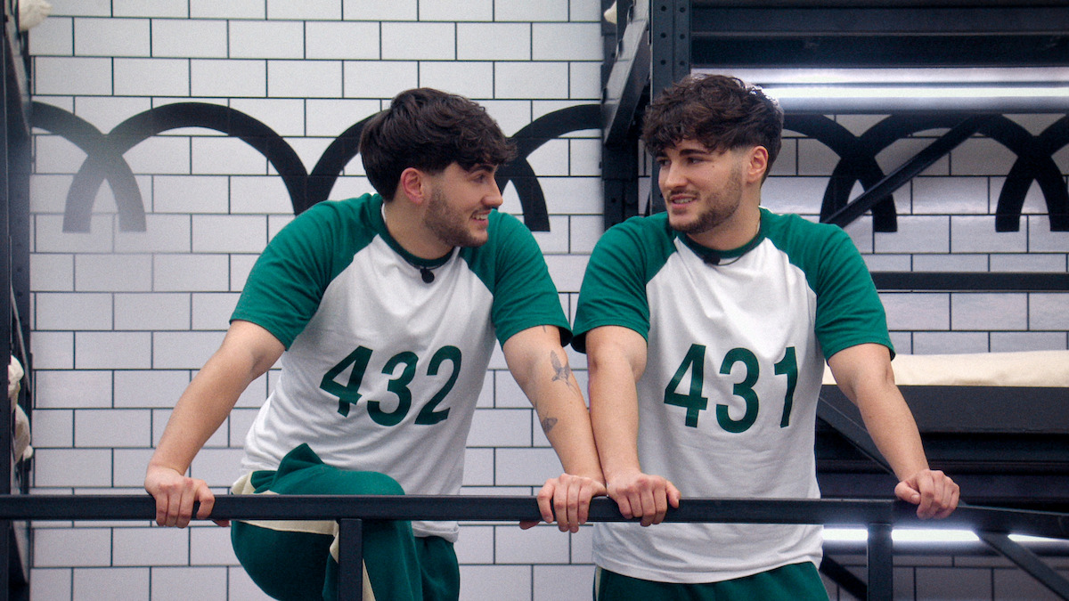 Two young men in matching green and white sports uniforms numbered 432 and 431 are talking and smiling on an indoor balcony with white tiled walls, giving a friendly and relaxed mood.