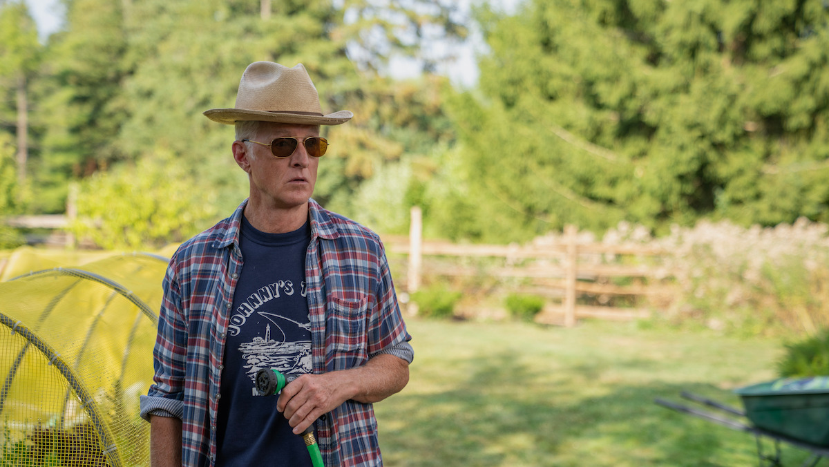 John Slattery as John wearing a hat and sunglasses, holding a garden hose in a sunny backyard garden with greenery, wooden fence, and trees in the background. Casual outdoor rural setting.