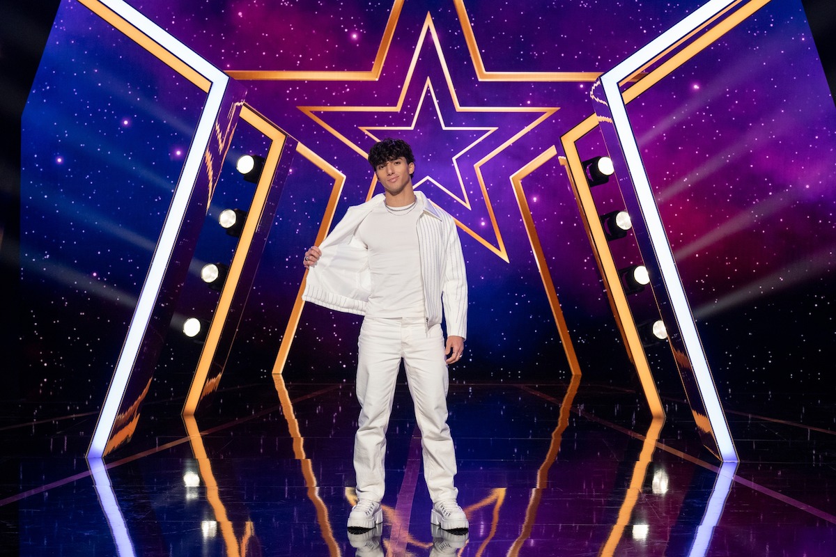 Issy Saadeh, vestida completamente de blanco, se encuentra con confianza en un escenario brillantemente iluminado con un telón de fondo con temática de estrellas, luces coloridas y un piso reflectante, creando un ambiente de espectáculo vibrante.