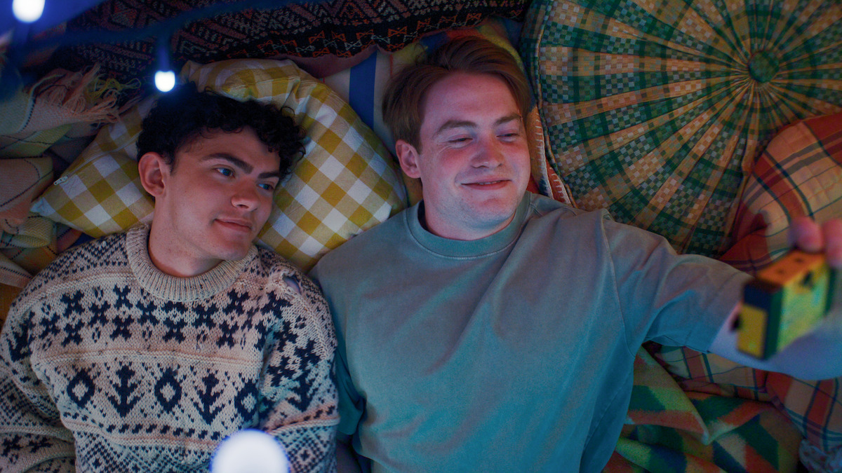 Two young people lying on colorful patterned blankets and cushions, one holding up a camera to take a photo, appear relaxed and happy in a cozy, softly lit indoor setting.
