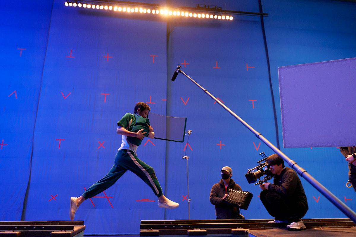 Actor in green tracksuit jumps across set with blue screen backdrop, surrounded by film crew, camera operator, and boom mic, indicating a behind-the-scenes film production environment.