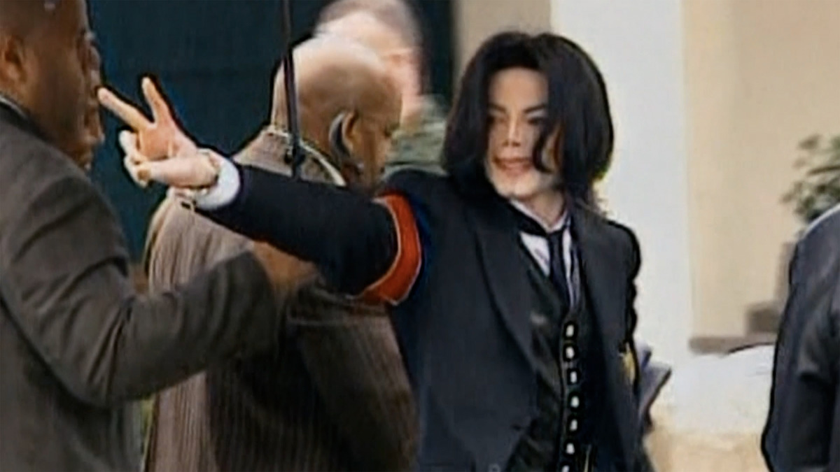 Person in a black suit with red armband making a peace sign outdoors, surrounded by several people in a public or official setting.