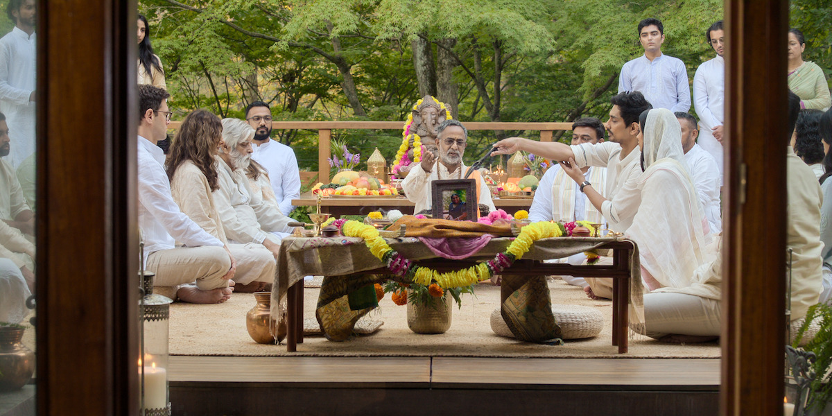 A group of people dressed in white participate in a traditional religious ceremony outdoors, seated around a decorated altar with offerings, food, flowers, and a portrait, surrounded by greenery and trees in the background.