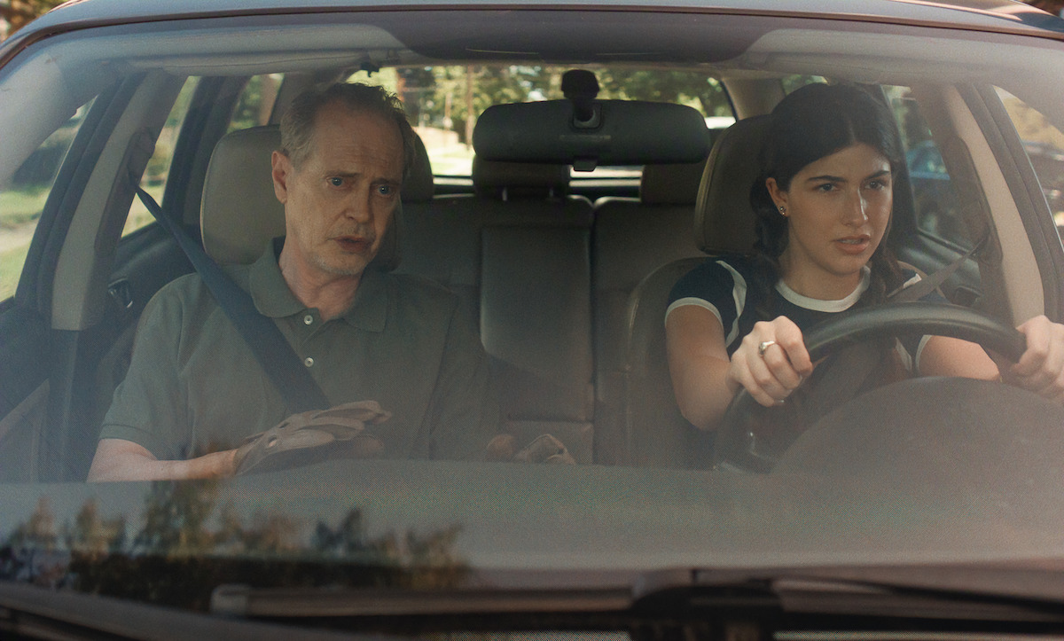 A teenage girl drives a car with an older man in the passenger seat; both look serious and focused, seen from the front windshield on a sunny day, likely during a driving lesson.