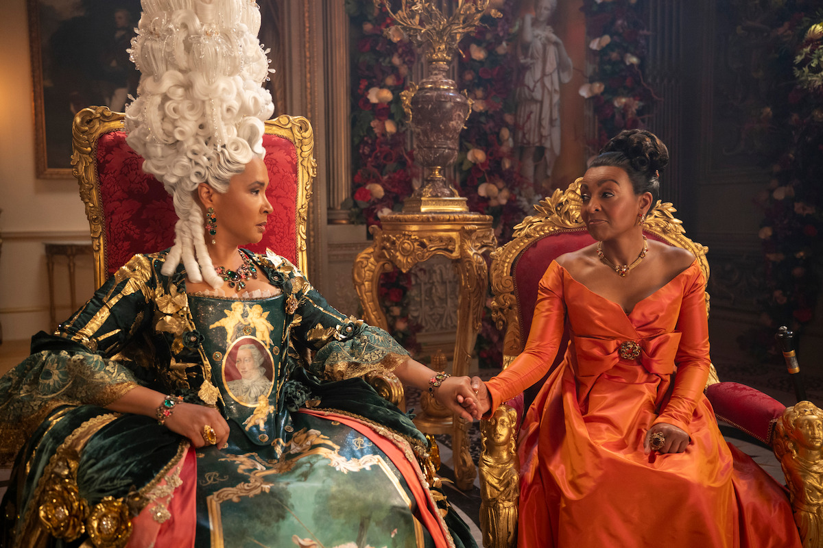 Two women in ornate historical dresses sit on golden thrones in a lavish, flower-filled room, holding hands and looking at each other, suggesting a close relationship and a royal or aristocratic setting.