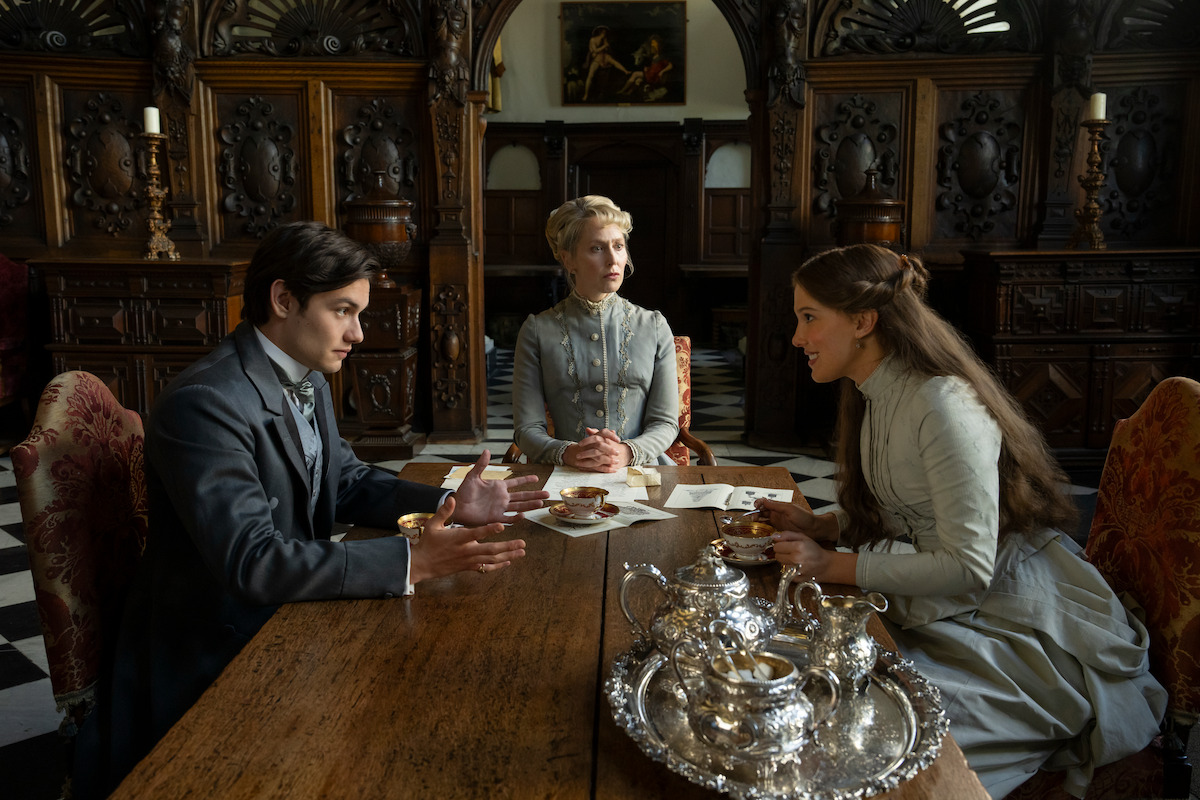 Three people in Victorian-era clothing sit around a wooden table with silver tea set in an ornate, wood-paneled room, engaged in conversation.