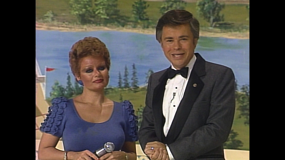 A woman in a blue dress holding a microphone stands beside a man in a tuxedo. Both are indoors with a painted scenic backdrop showing trees and water, suggesting a formal event or television show setting.