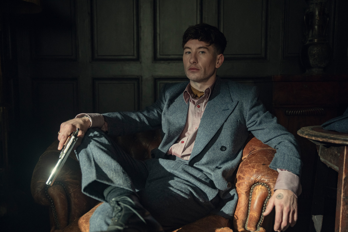 Man in a gray suit sitting in a vintage leather armchair, holding a gun, in a dimly lit room with dark paneled walls, creating a tense and dramatic atmosphere.