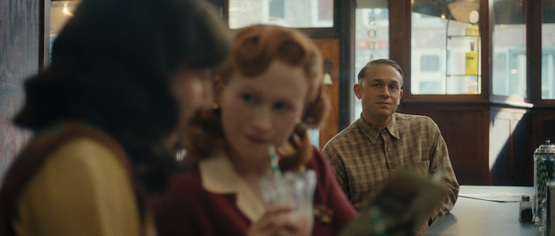 A man in a plaid shirt sits alone at a diner counter, looking at two women in the foreground, one of whom is sipping a drink with a straw.