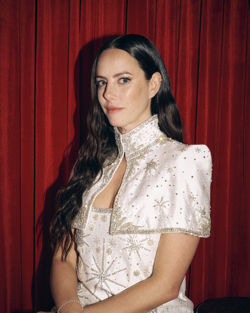 Woman with long dark hair in a white, embellished dress poses in front of red velvet curtains, looking at the camera in a formal portrait setting.