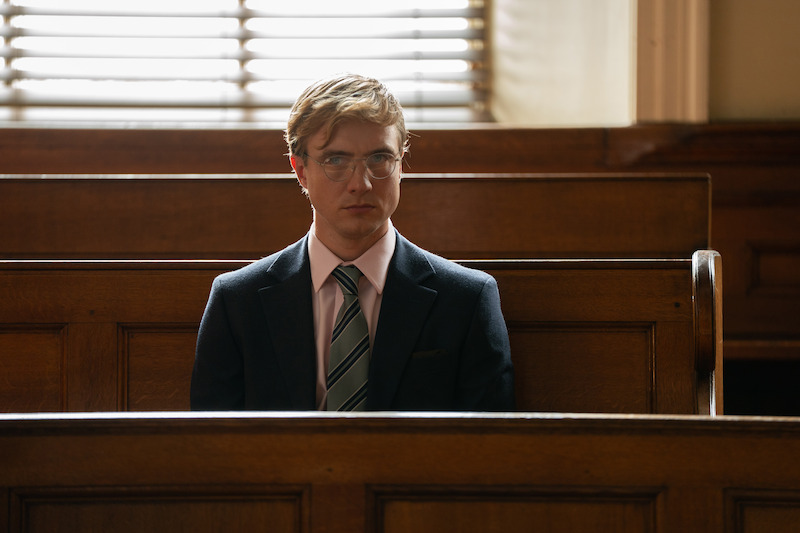 A man in a suit and glasses sits alone on a wooden bench in a courtroom, with sunlight streaming through window blinds behind him.