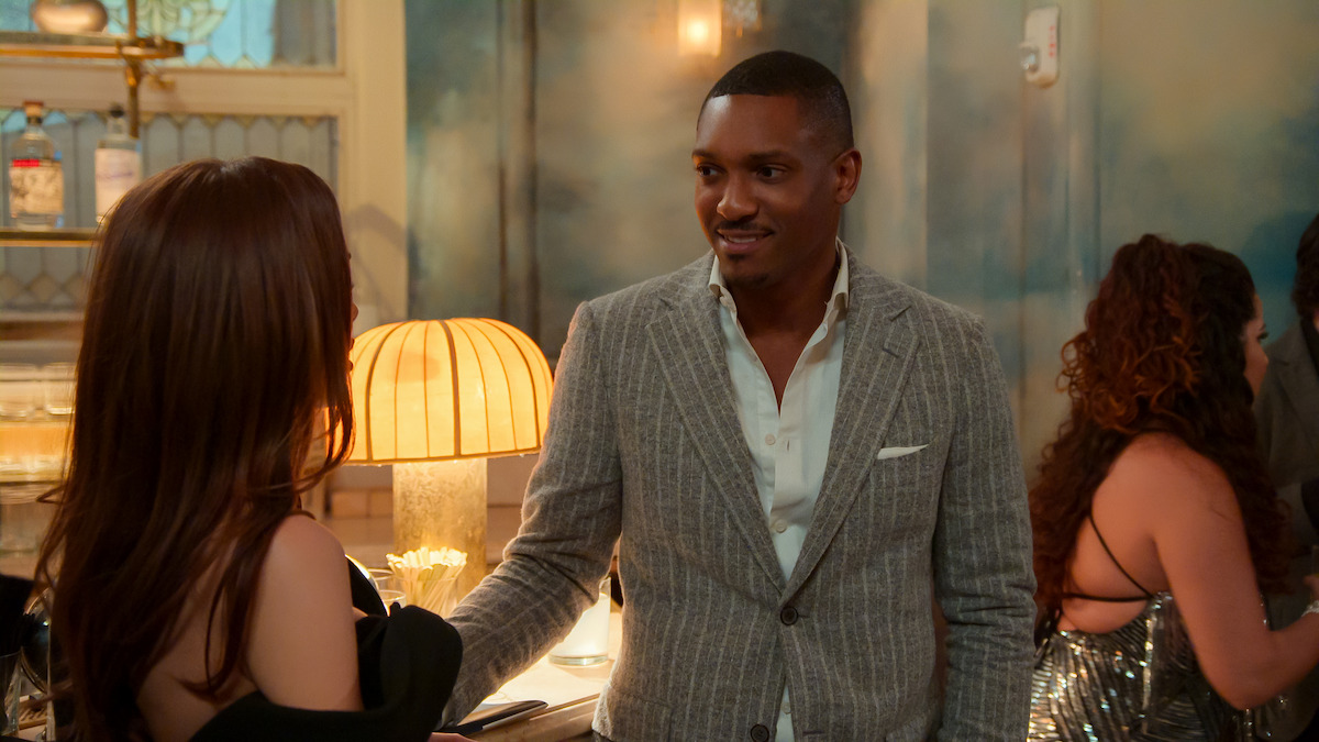 A well-dressed man smiles while conversing with a woman at an elegant indoor party; warm lighting and stylish decor create a sophisticated, relaxed atmosphere.