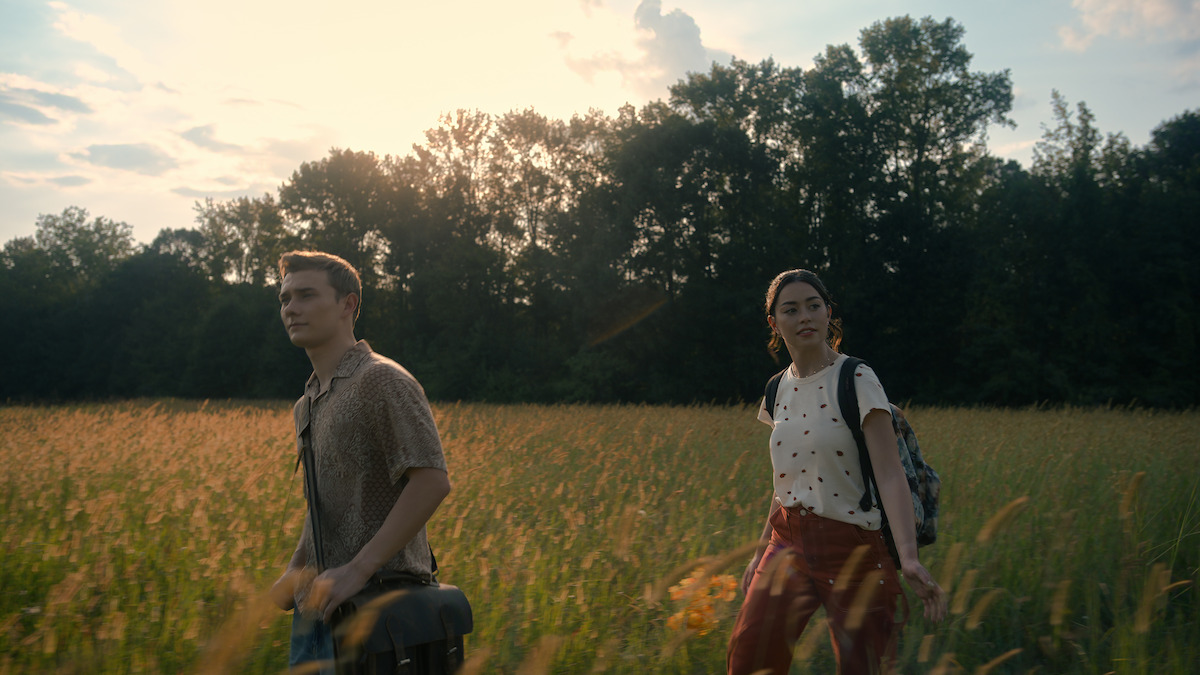 Two people walk through a sunlit grassy field with trees in the background. The sky is partly cloudy and the setting suggests late afternoon or early evening. Both individuals carry bags and appear to be exploring or hiking.