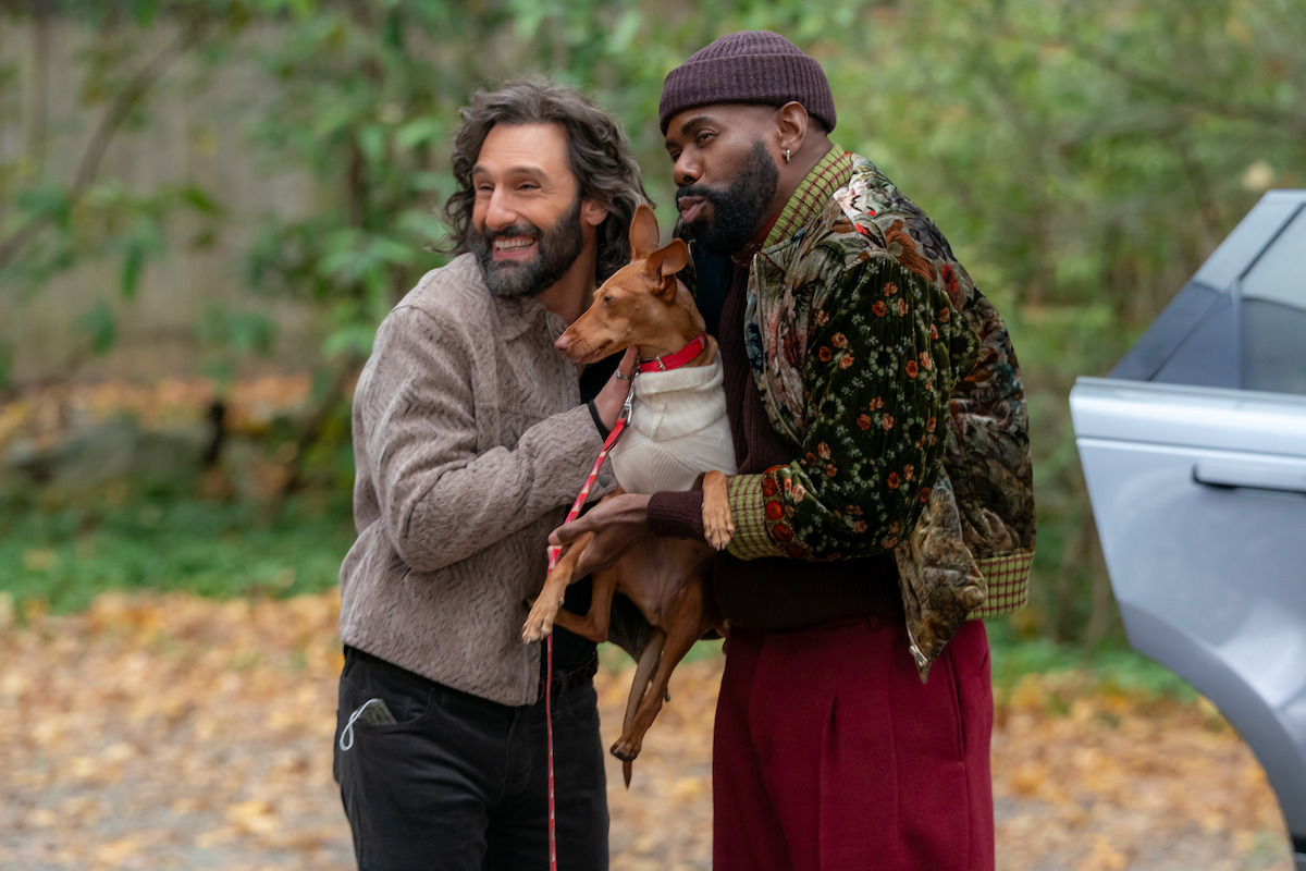 Two men outdoors standing close together, one holding a small brown dog on a leash. They are dressed warmly, surrounded by greenery and autumn leaves, and next to a silver car.