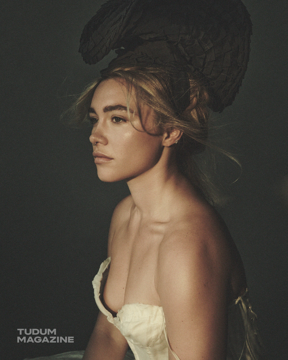 Woman in a dramatic feathered headpiece and strapless dress poses in profile against a dark, moody background, soft lighting highlighting her face and shoulders. "Tudum Magazine" logo appears in the lower left corner.