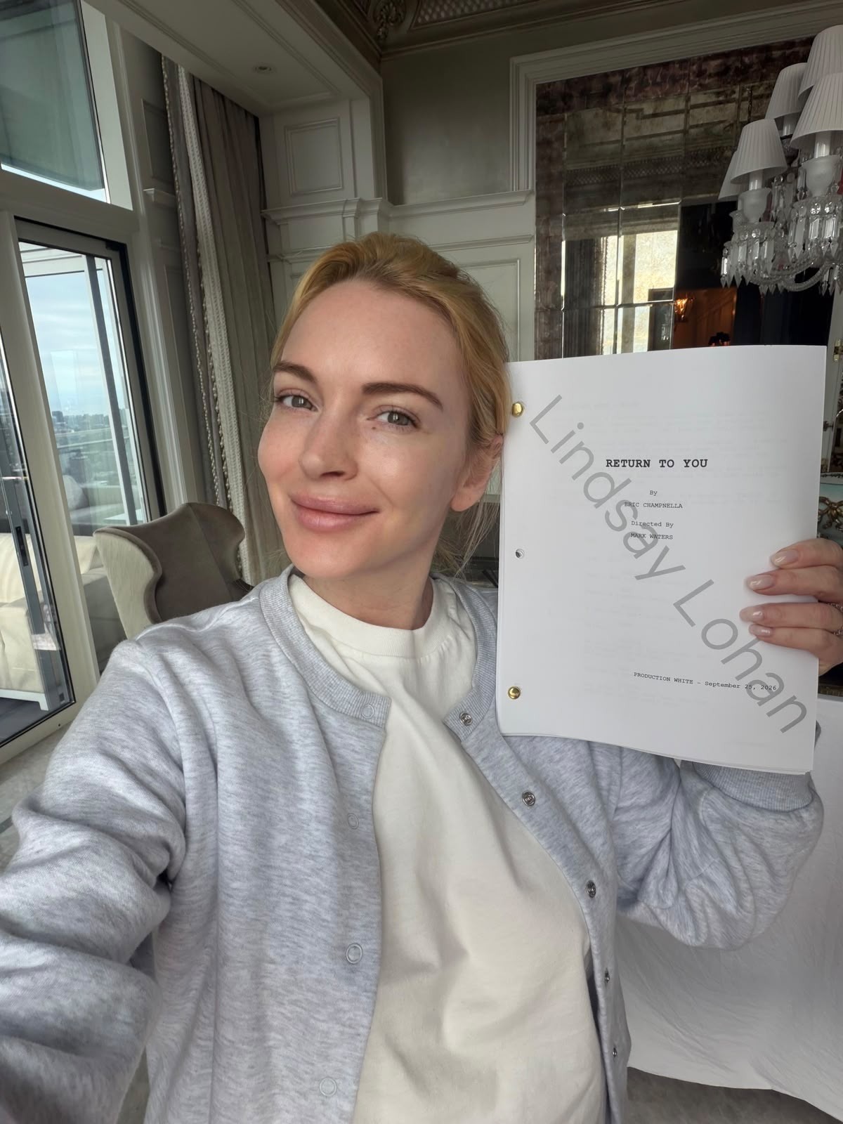 Woman taking a selfie in a bright, elegant room, holding up a script titled "Return to You." She is smiling, with daylight coming through large windows and a chandelier above.
