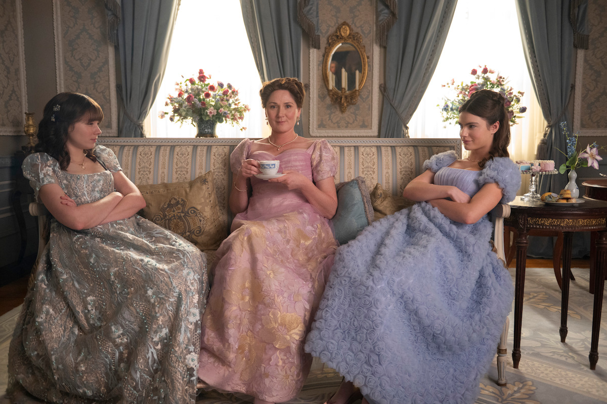 Three women in elegant period dresses sit on a vintage sofa in a richly decorated room, two looking displeased with arms crossed, one smiling with a teacup, creating a formal yet tense atmosphere.