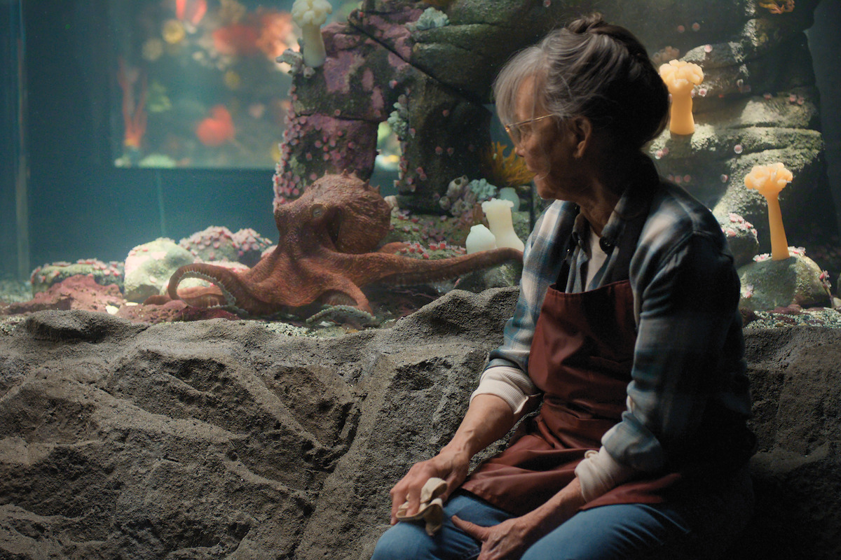 Sally Field as Tova in an apron sitting in front of a rocky aquarium tank, looking at an octopus inside. The tank features colorful coral, rocks, and soft lighting, suggesting an indoor aquarium environment.