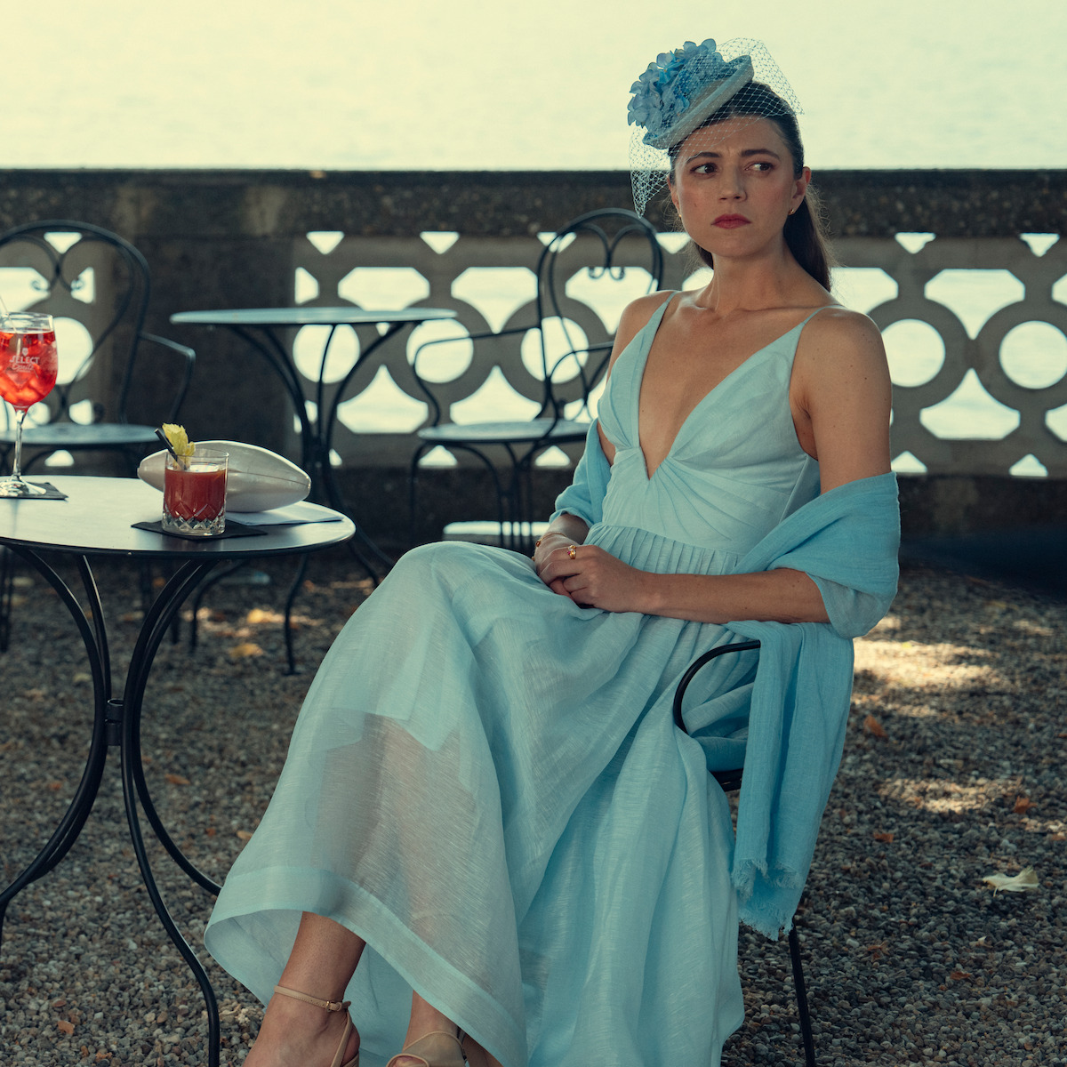 Woman in light blue dress and hat sits at an outdoor café table near a lake, with drinks and a bowl nearby, stone balustrade in the background, elegant and serene summer setting.