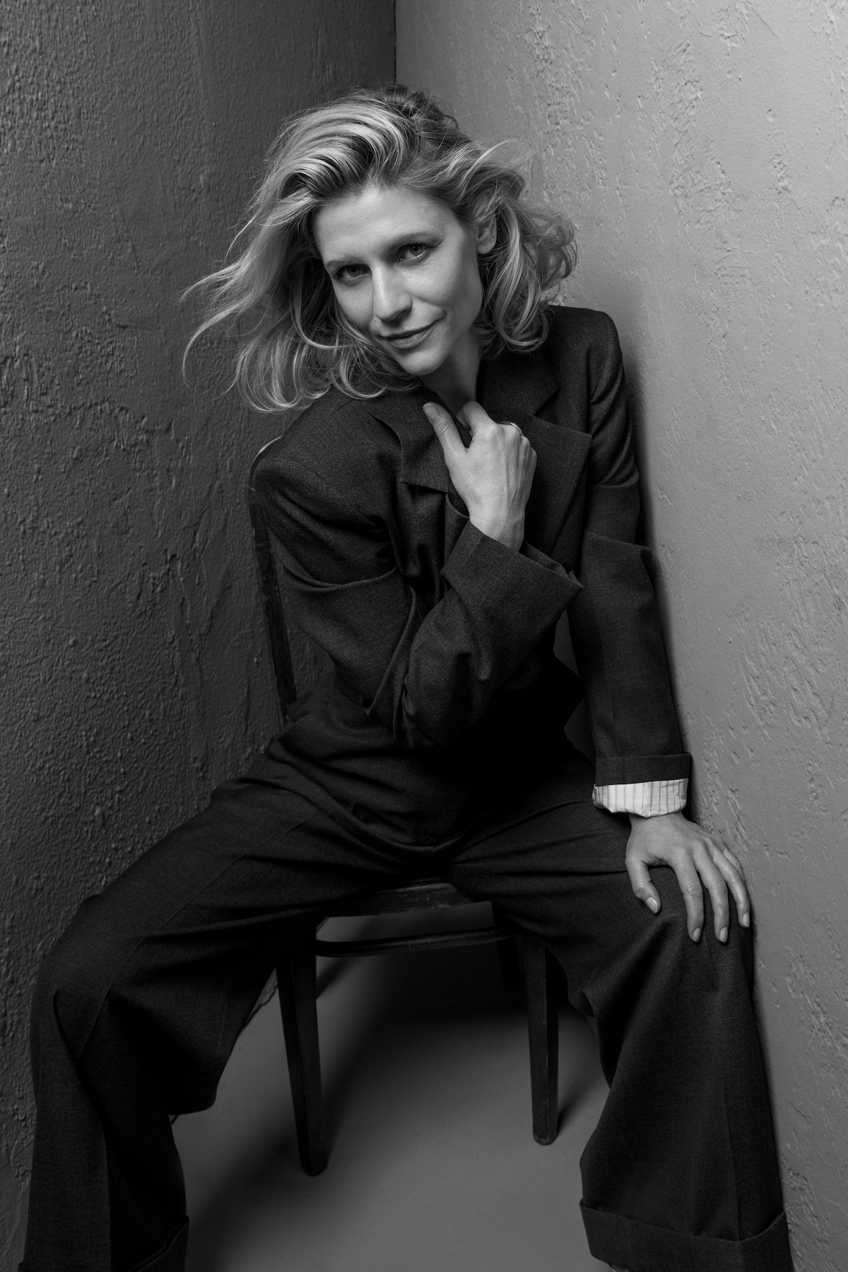 Woman in a loose suit sits confidently on a chair in a narrow, textured-walled corner. Monochrome lighting adds a dramatic, edgy, and stylish mood to the portrait.
