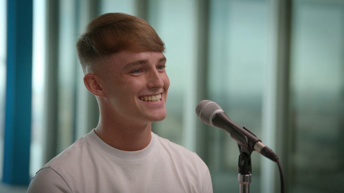 Young man smiling while sitting in front of a microphone in a modern, bright room with large windows, creating a cheerful and relaxed atmosphere.