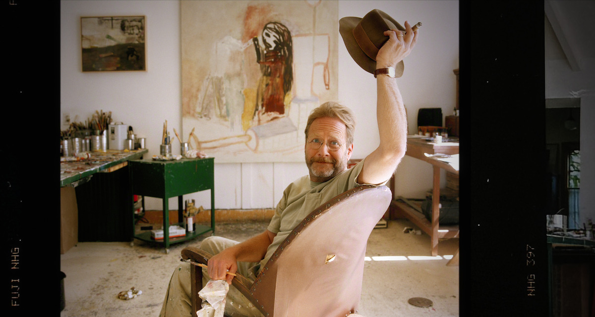 Man sitting in an artist’s studio, holding up a hat and smiling. The room has art supplies, canvases, and a large painting in the background, giving a creative and relaxed atmosphere.