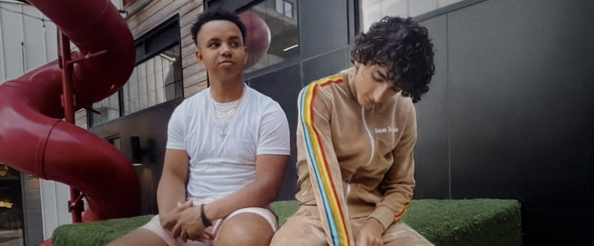 Two people sit on a green bench outdoors in an urban area, with one wearing a white t-shirt and shorts and the other in a tan tracksuit with rainbow stripes. A large red tube slide and modern buildings are visible in the background.Two people sit on a green bench outdoors in an urban area, with one wearing a white t-shirt and shorts and the other in a tan tracksuit with rainbow stripes. A large red tube slide and modern buildings are visible in the background.