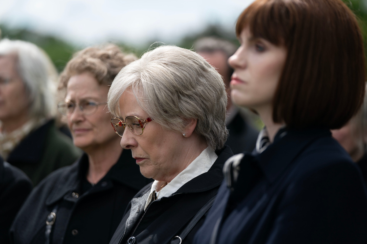 Several somberly dressed women stand closely together outdoors, appearing solemn and reflective, likely attending a serious event such as a funeral or memorial service, with a blurred natural background.