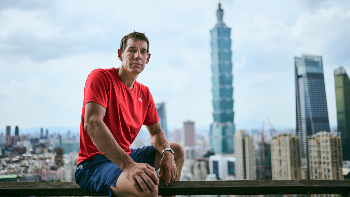 Man in a red shirt sitting on a rooftop railing overlooking a city skyline with tall buildings and the Taipei 101 skyscraper in the background on a cloudy day.