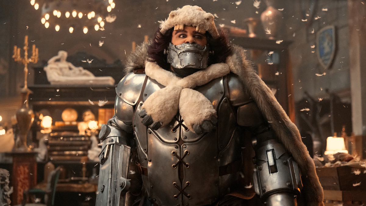 Rob Colletti as Wapol, in heavy metal armor with fur accents stands indoors, surrounded by snowflakes in a room with medieval decor, chandeliers, and warm lighting, creating a dramatic and fantasy-themed atmosphere.