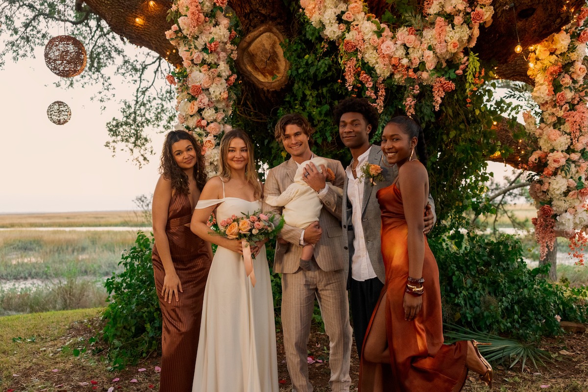 Group posing at an outdoor wedding under a large tree decorated with pink and white flowers, smiling and dressed in formal attire, with scenic landscape in the background.