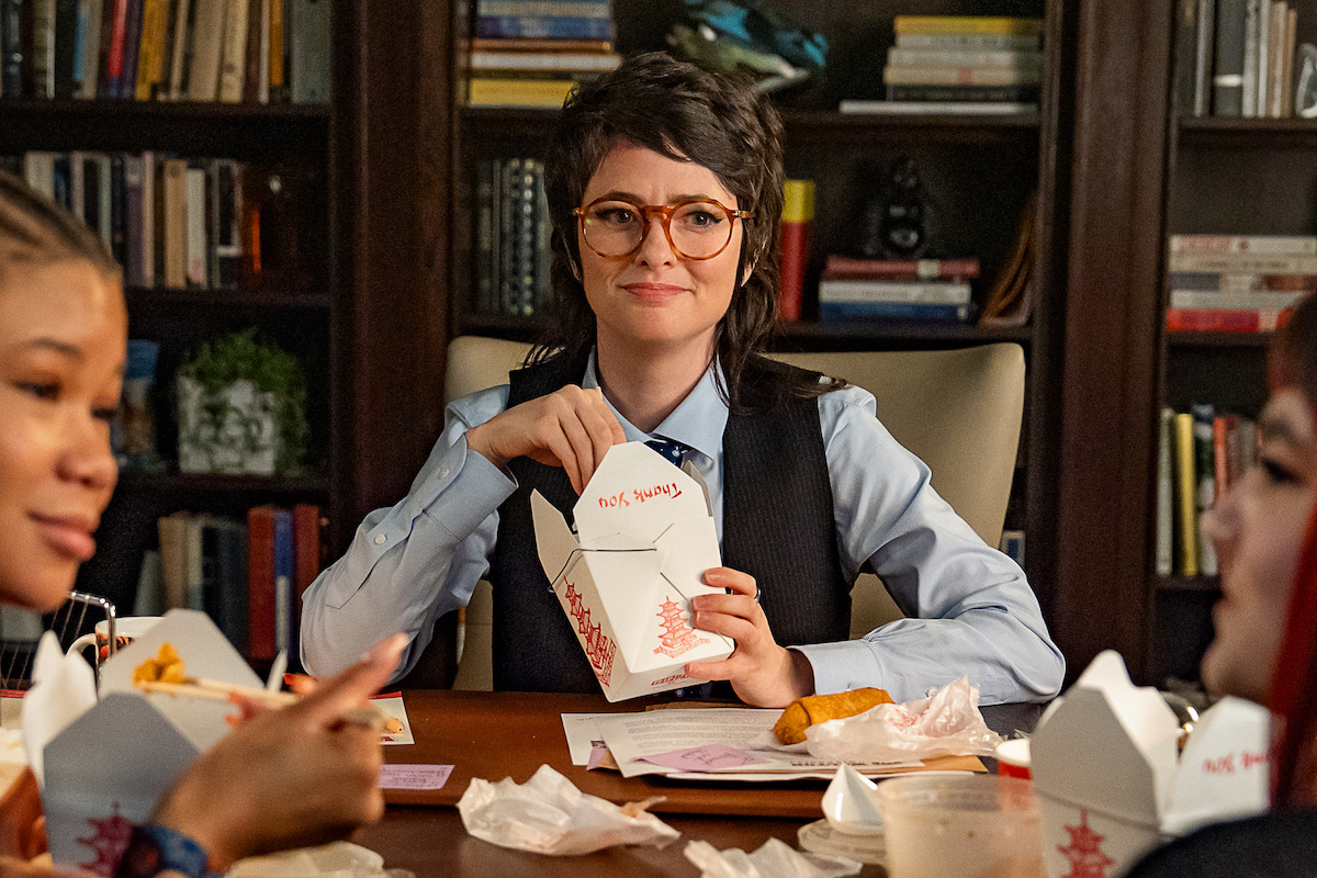 Person in glasses enjoying takeout food with friends at a table, surrounded by open Chinese food containers and drinks, in a cozy room with bookshelves filled with books in the background.