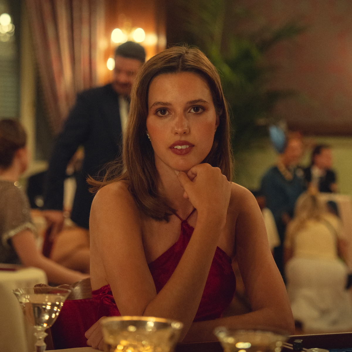 A woman in a red dress sits at a dinner table in an elegant, dimly lit restaurant with other people socializing in the background, gold-rimmed glasses on the table, soft lighting and a formal, vintage atmosphere.