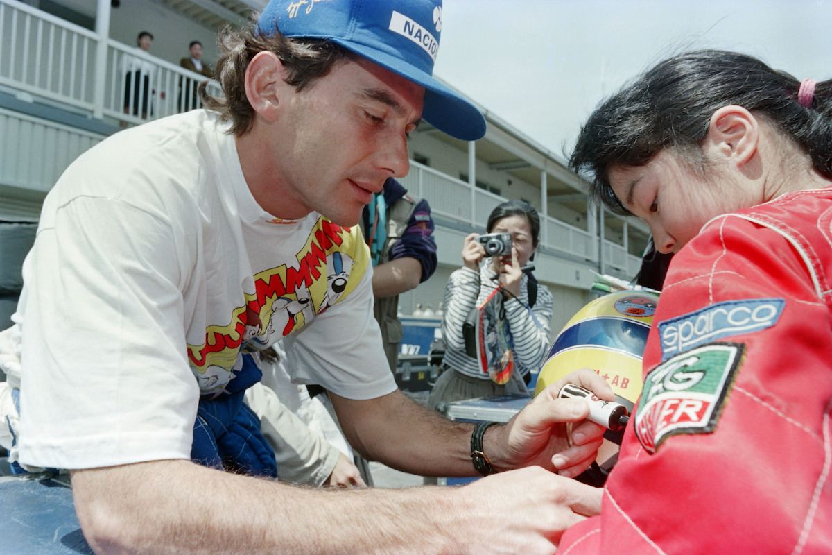 Senna, wearing a white tshirt and blue hat bends down to sign the racing suit of a young female fan.