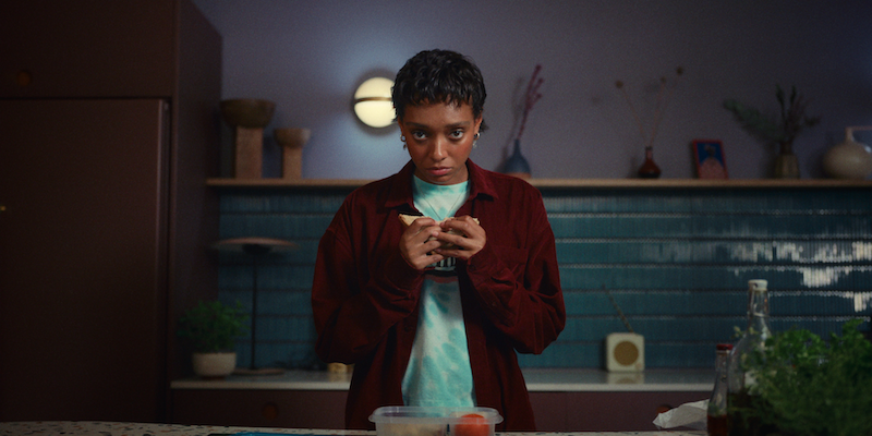 Sophie Wilde as Mia stands in a kitchen holding a piece of bread in Season 1 of 'Everything Now.'