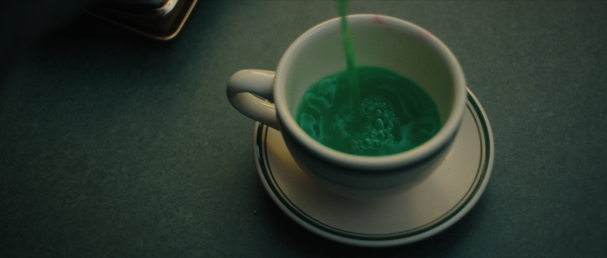 A close-up of a white cup on a saucer placed on a green surface, being filled with a bright green liquid, creating ripples inside the cup. The lighting is soft and moody, giving a calm and mysterious atmosphere.